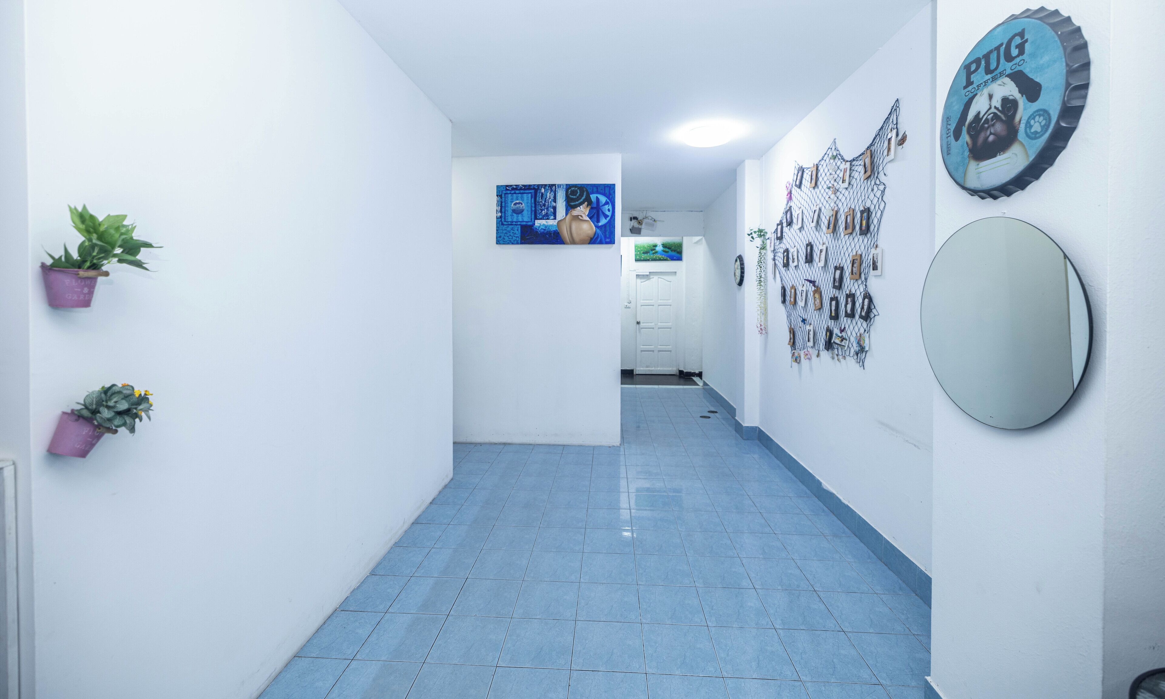 Un pasillo con suelo de baldosas, paredes blancas y una puerta al final. Hay dos macetas con plantas en la pared izquierda y un cuadro enmarcado sobre la puerta.
