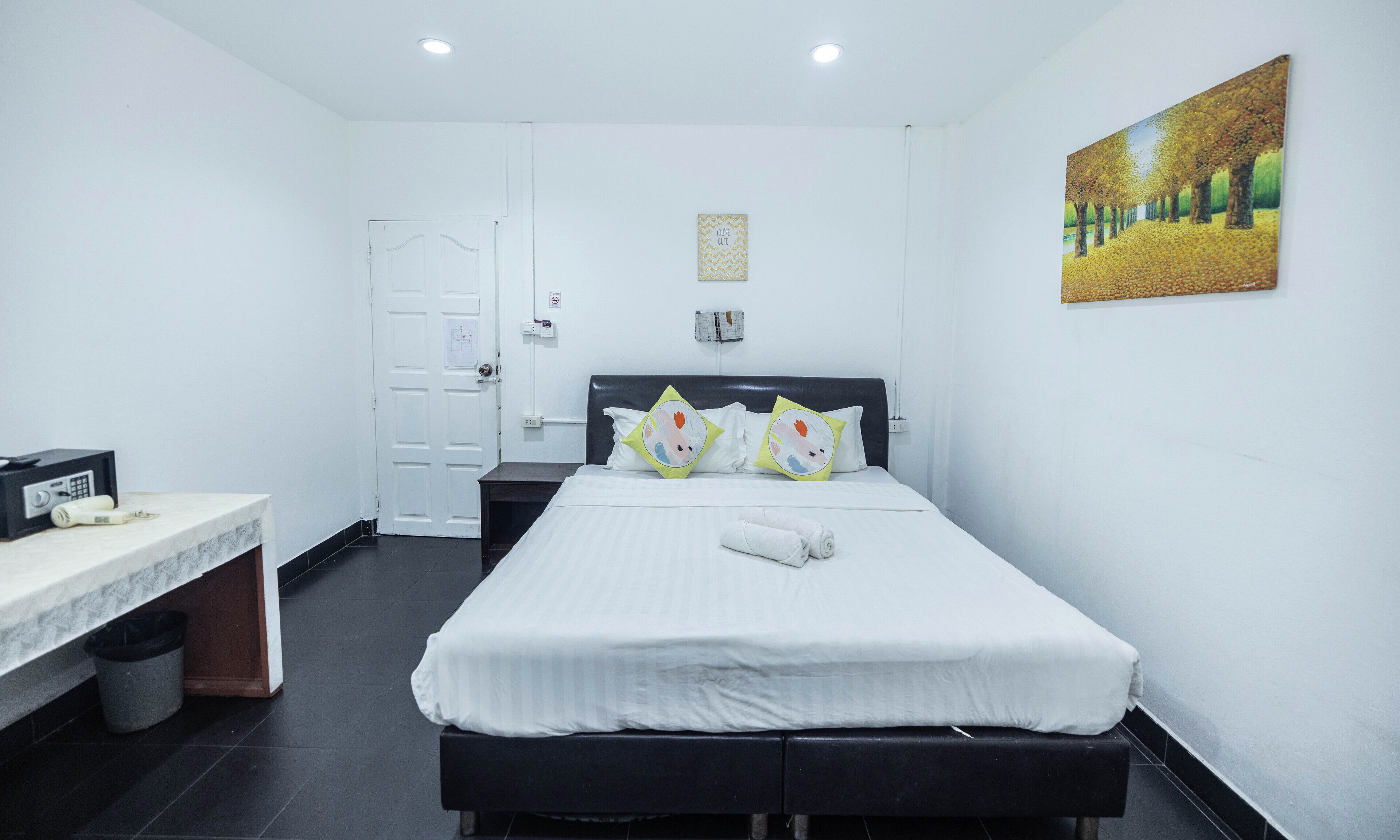 A hotel room with a bed, a desk, a chair, a trash bin, and a painting on the wall.