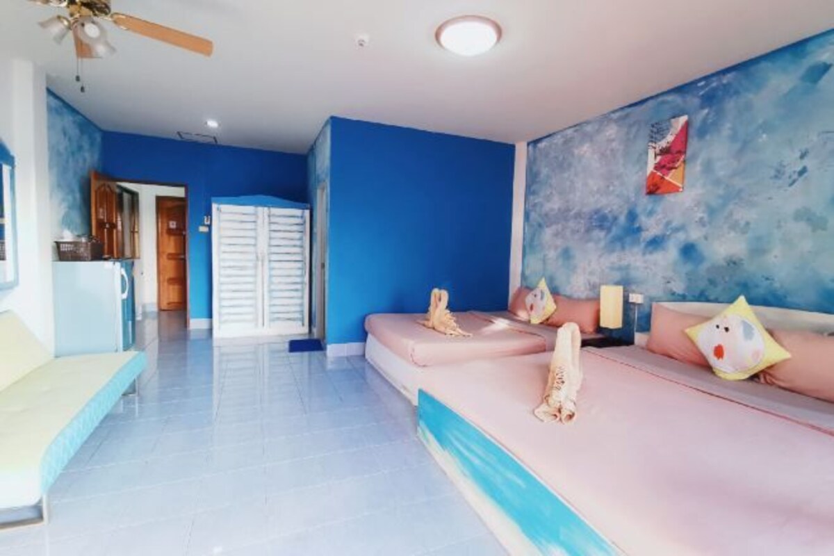 Una habitación con pared azul, cama con ropa de cama rosa, un sofá, un ventilador de techo y una puerta que da a otra habitación.