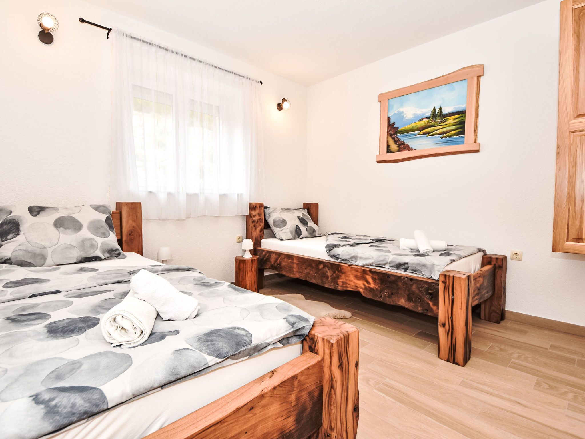 A bedroom with two wooden beds, a painting on the wall, and a window with sheer curtains.