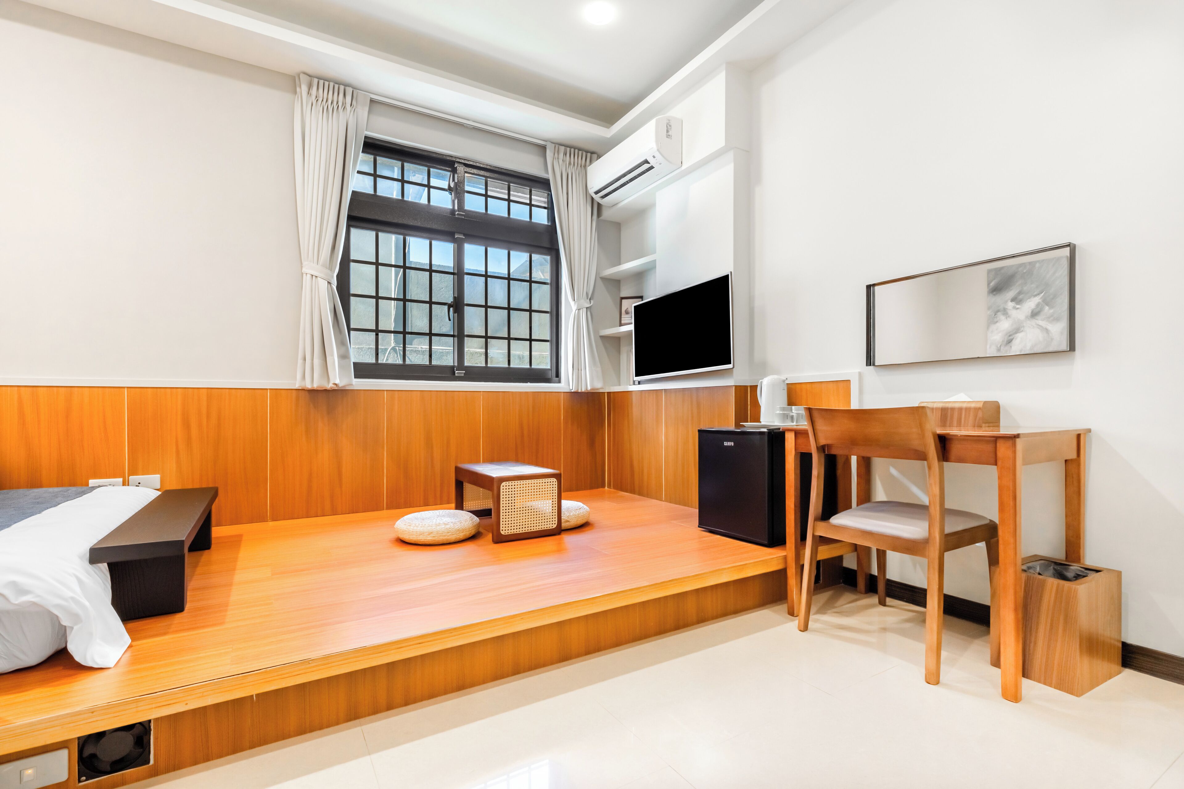 Una habitación de hotel moderna con un banco de madera, una mesita, una silla, un televisor y un ventanal con cortinas.