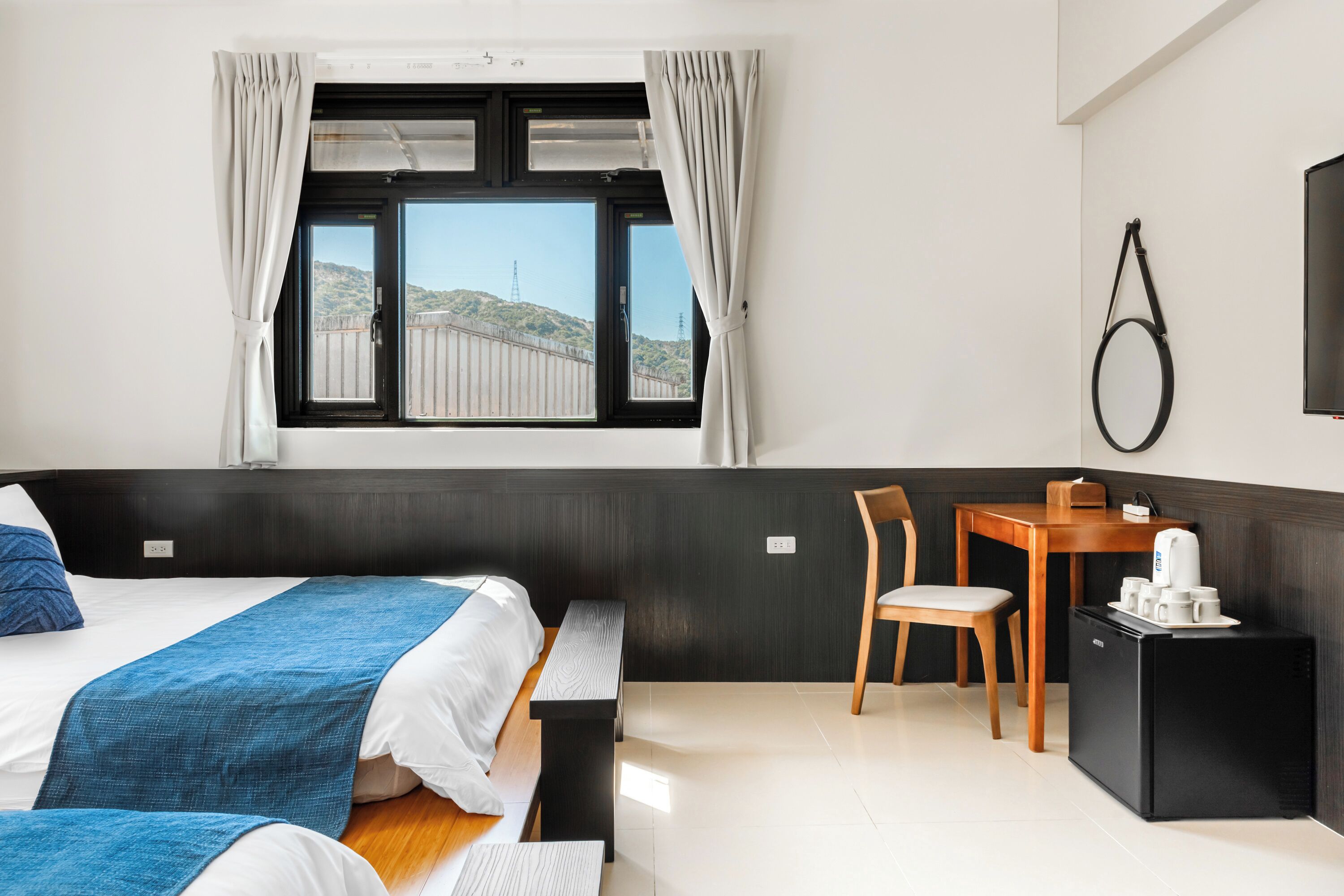 A hotel room with two beds, a desk, a chair, a small refrigerator, and a window with curtains.