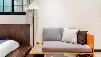 A compact room with a wooden bed, a grey sofa with cushions, a glass table, and a floor lamp.