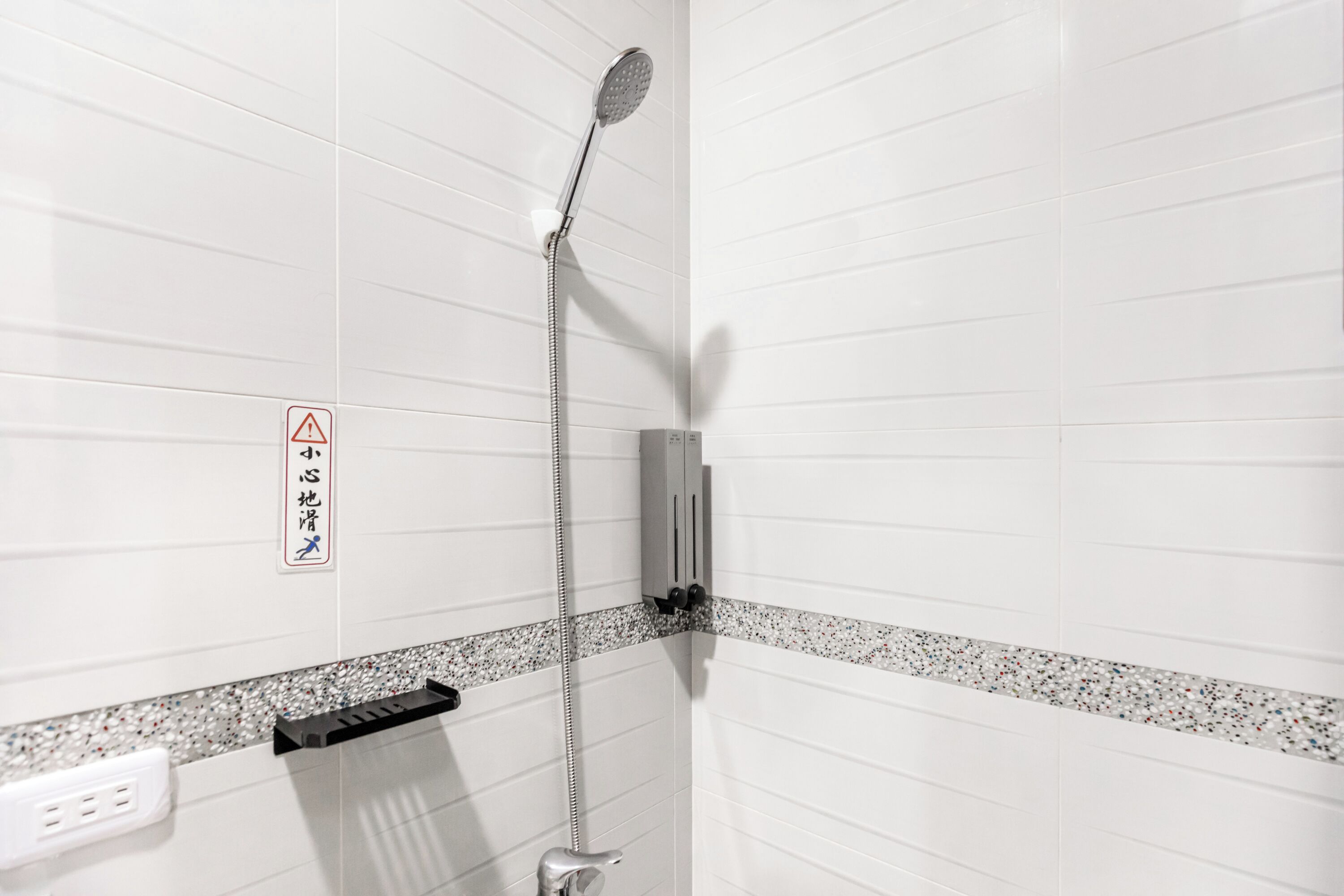 Un espace douche avec des murs recouverts de carreaux blancs, une pomme de douche, un panneau de commande et un panneau d’avertissement.