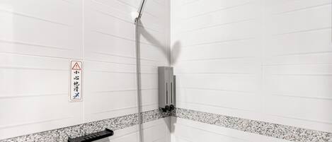 Un espace douche avec des murs recouverts de carreaux blancs, une pomme de douche, un panneau de commande et un panneau d’avertissement.