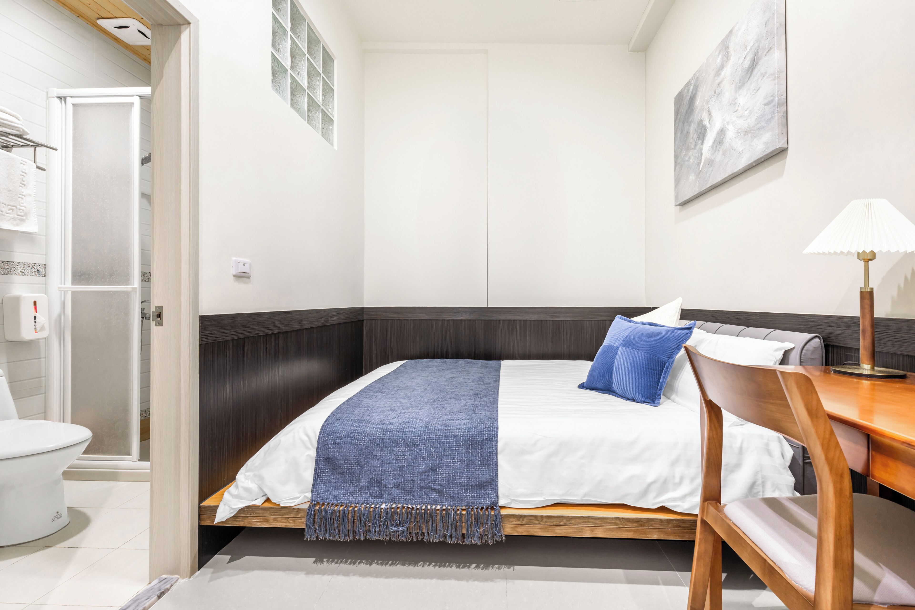 A hotel room with a bed, a wooden desk, a chair, a lamp, and a bathroom visible through an open door.