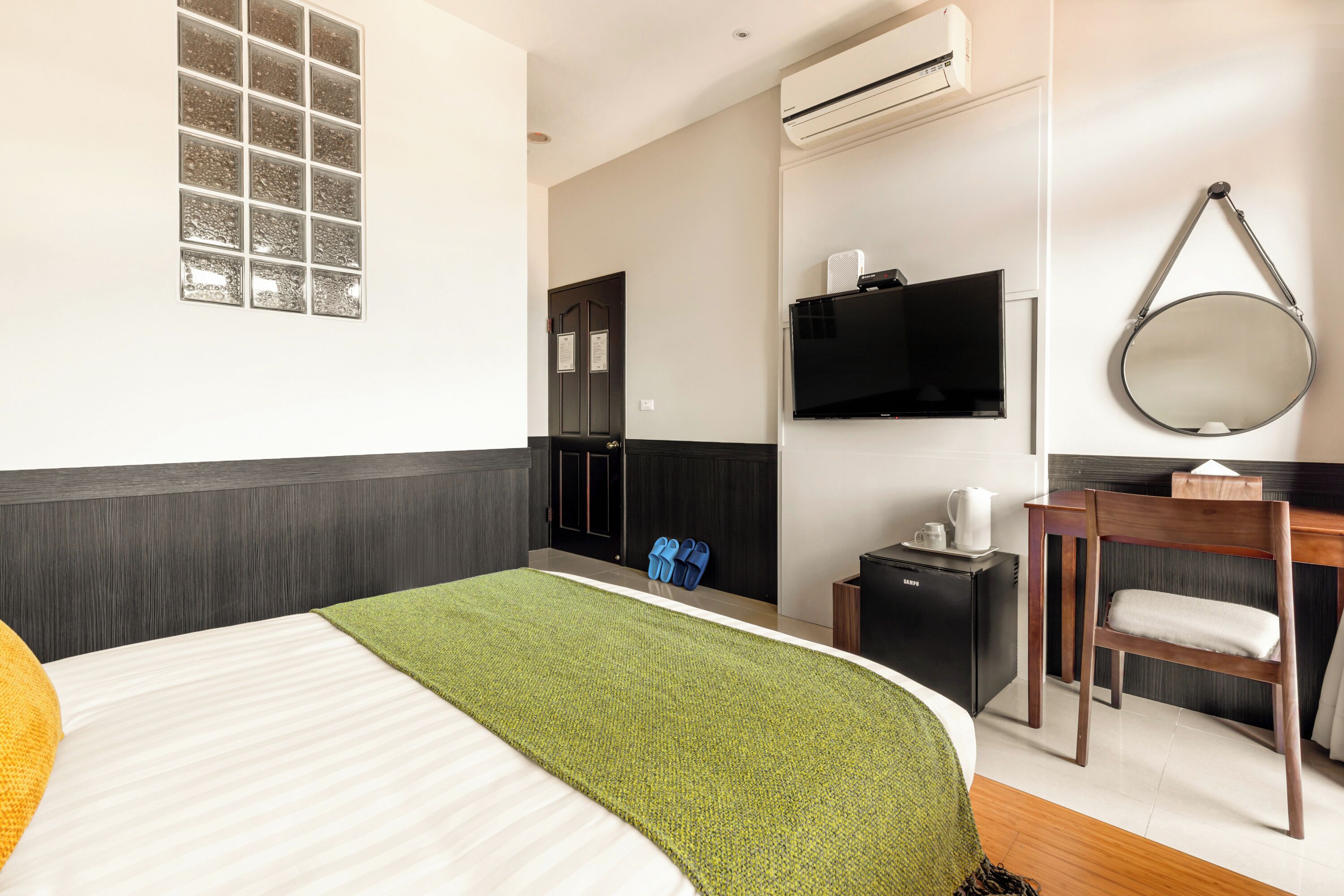 A hotel room with a bed, a green throw, a TV, a mirror, a desk, a chair, and a small cabinet.
