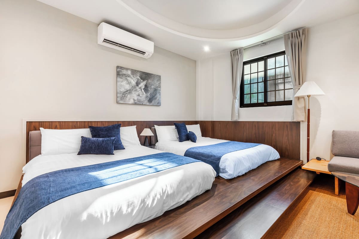 Habitació d'hotel amb dos llits, una finestra gran amb cortines, aire condicionat fixat a la paret i un quadre.