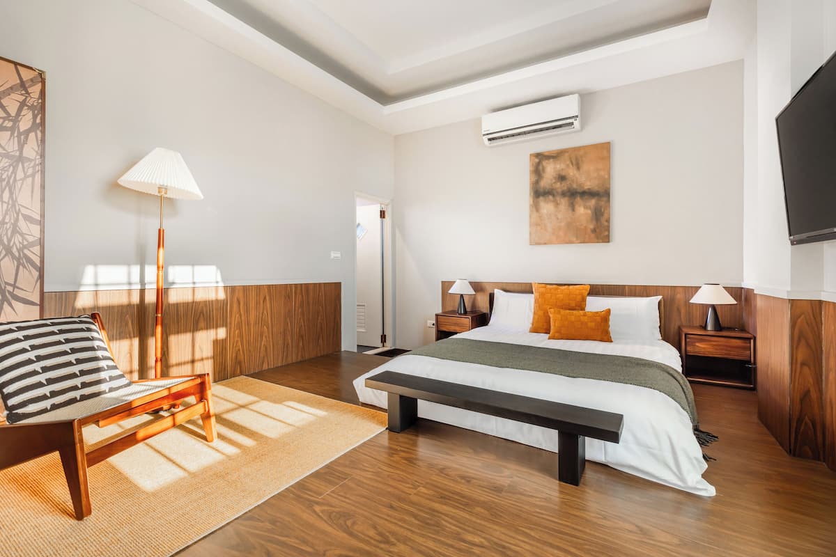 Habitació d'hotel moderna amb sòl de fusta, un llit gran amb capçalera, una taula de nit amb una làmpada, una panxa, una cadira i una televisió penjada a la paret.