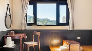 A room with a wooden table, a chair, a small desk with a kettle, a speaker, and a view of a mountain through a window. - Harmony Easy Stay (New Taipei City)