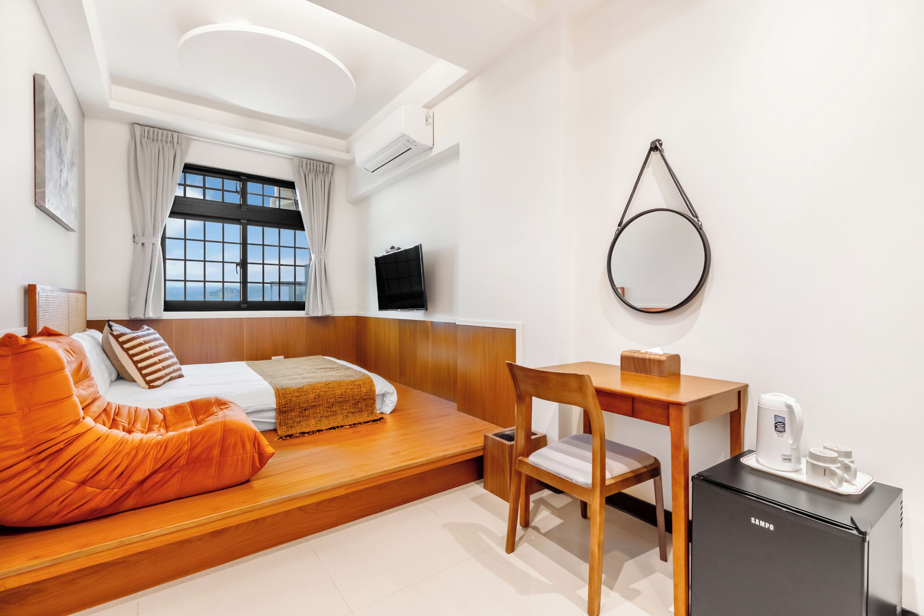 A modern hotel room with a bed, orange bean bag, wooden desk, and a small refrigerator.