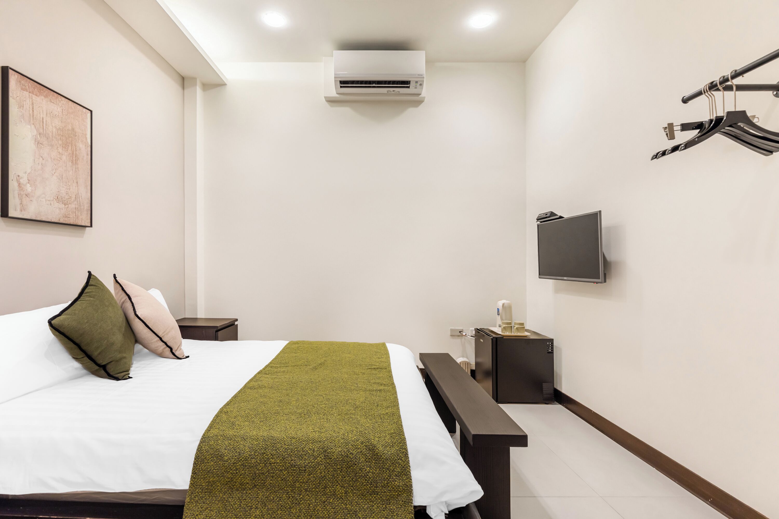 A hotel room with a bed, a flat-screen TV, an air conditioner, a bench, and a coat rack.