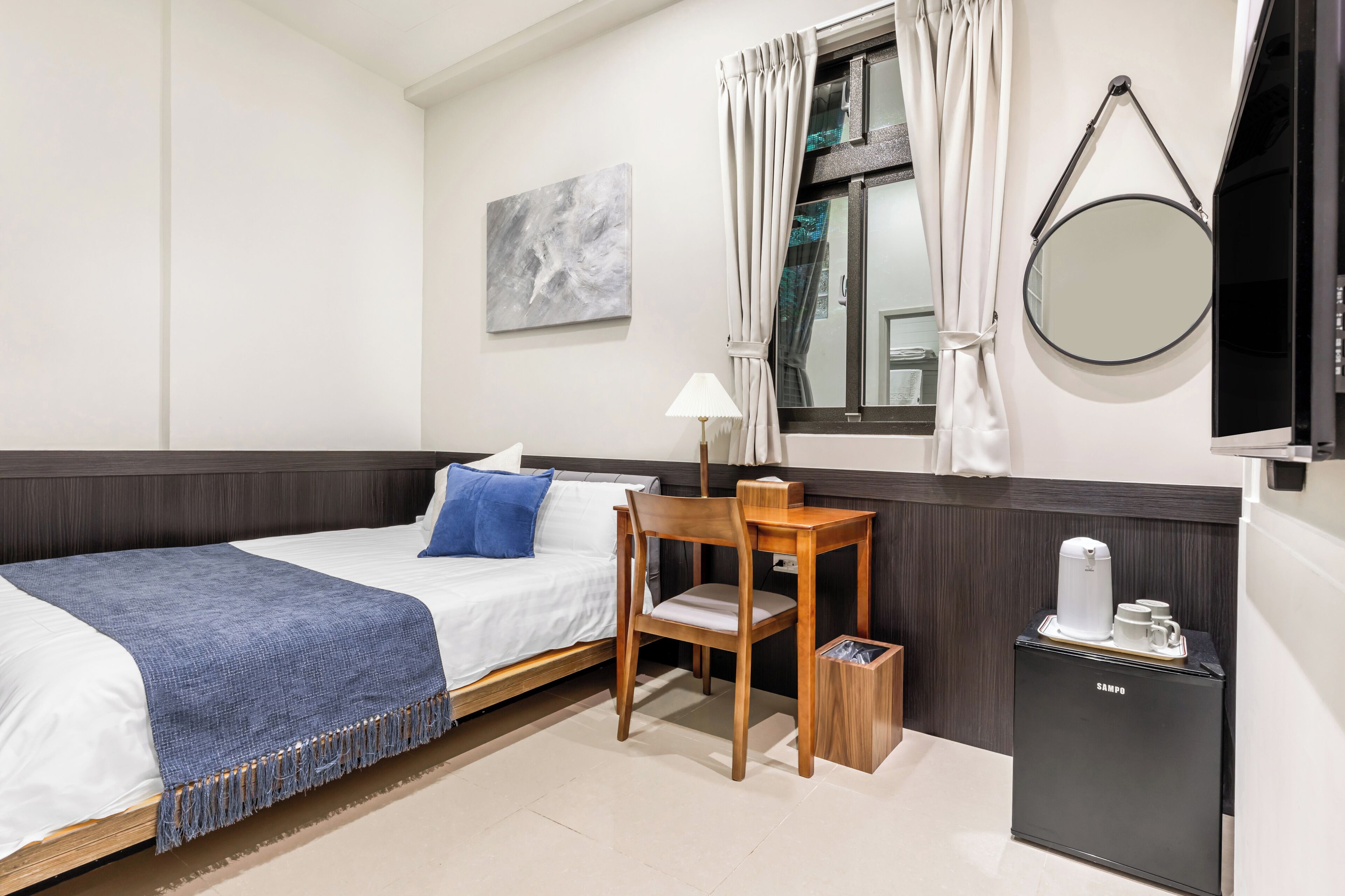 A hotel room with a bed, a desk, a chair, a small cabinet, a mirror, and a window with curtains.