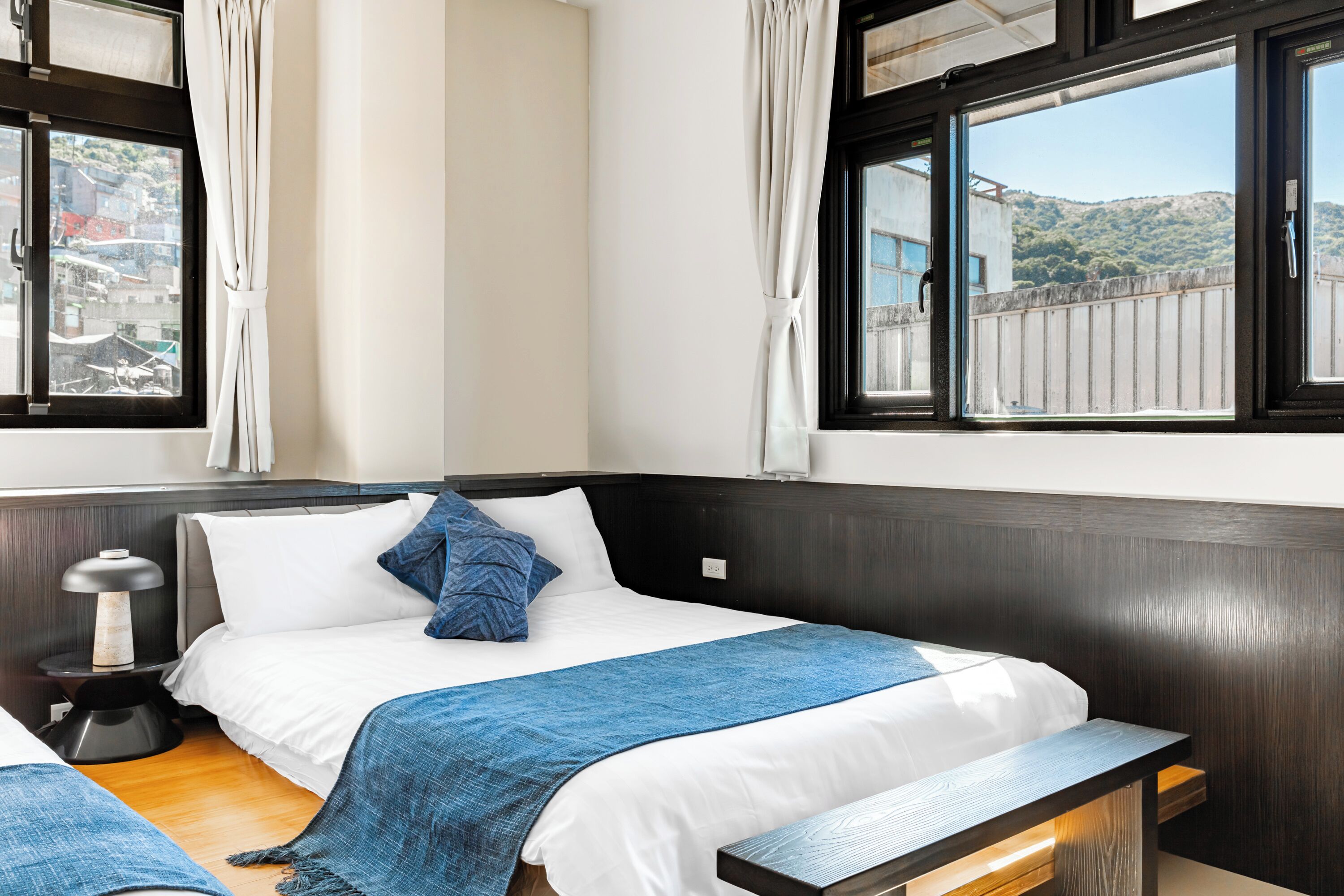 A hotel room with a bed, bedside table, lamp, and a view of a hillside.