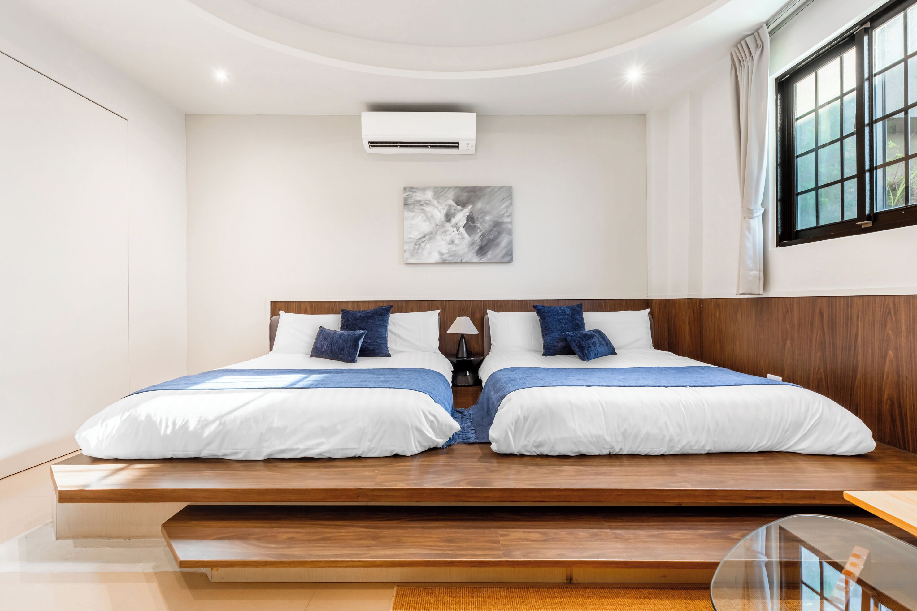 Quarto moderno com cama central, mesa de cabeceira pequena, ar condicionado fixado na parede e janela com cortinas.