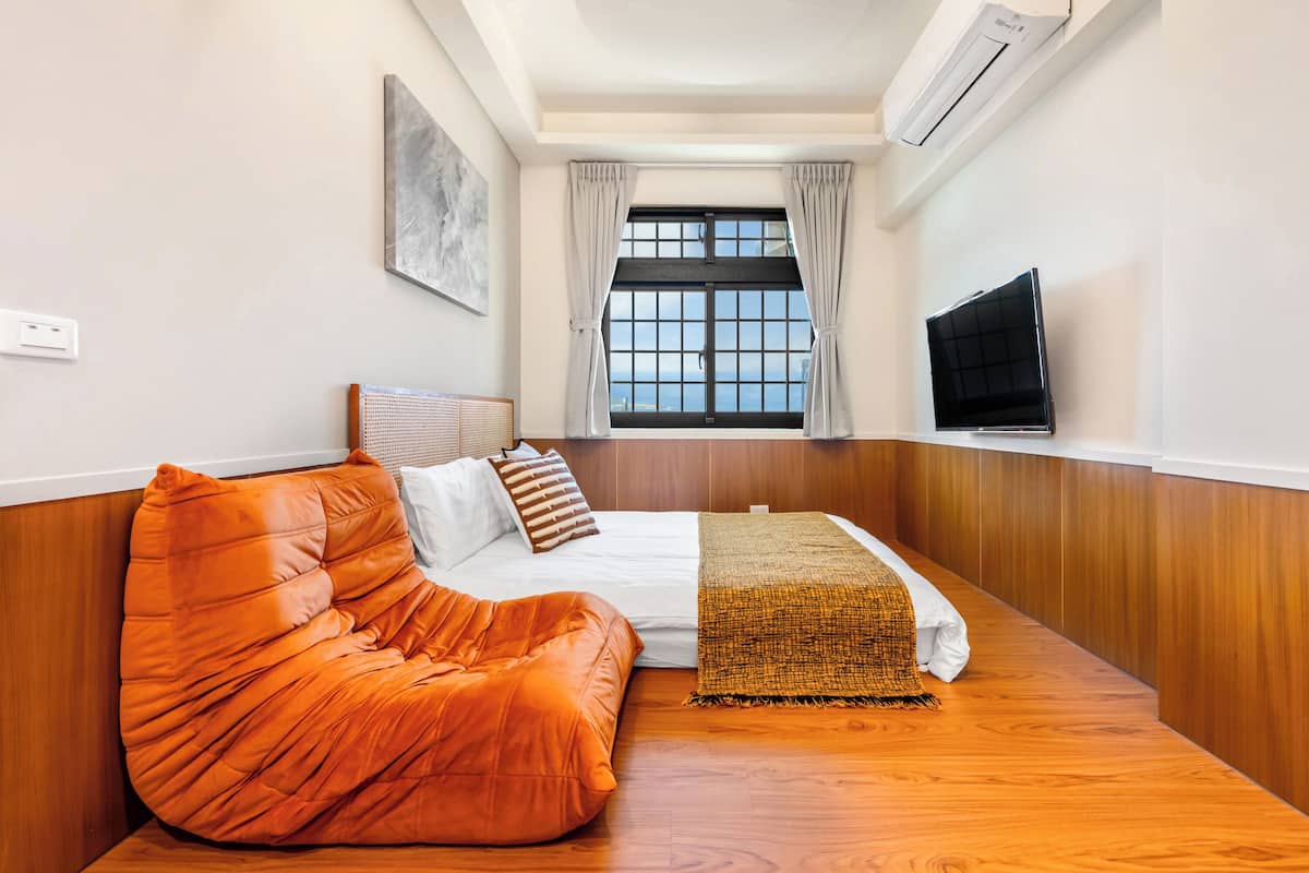 Habitació amb un llit, una butaca de sacs taronja, un televisor, una finestra amb cortines i aire condicionat.