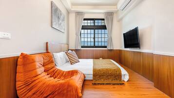 A room with a bed, orange bean bag, a TV, a window with curtains, and an air conditioner.