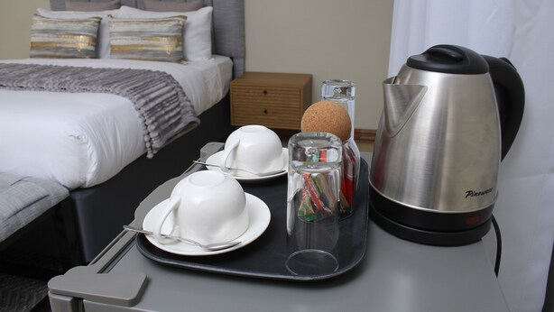 A neatly made breakfast tray with white cups, a glass of juice, and a silver kettle in a hotel room. - The Oak Tree House (Gaborone)