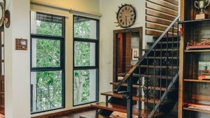 A room with a wooden staircase, large windows with a view of greenery, and a wall clock. - Khaoyai Valley 2 (Pak Chong)