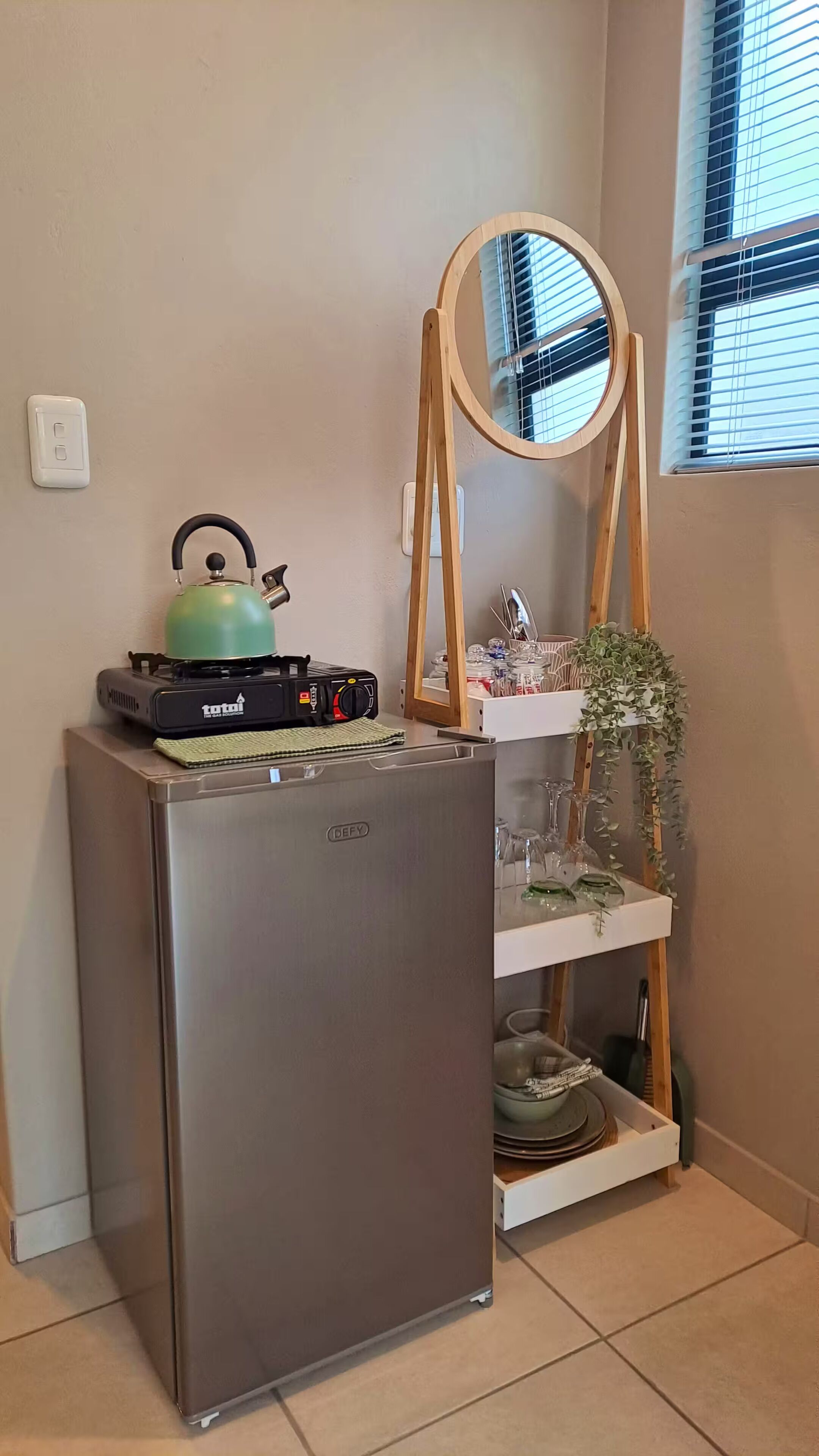 A compact kitchen with a built-in refrigerator, a kettle, a portable gas stove, and a wooden shelf with a mirror and glassware.