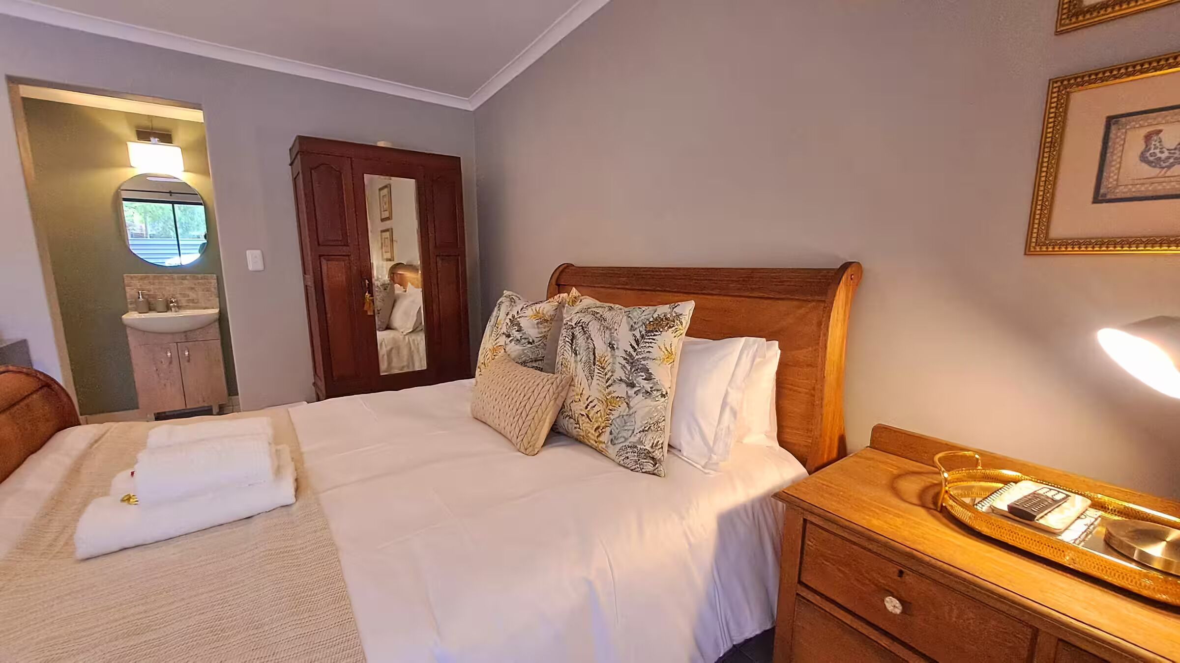Un lit bien fait, avec du linge de lit blanc et des oreillers à motifs floraux, une table de chevet en bois avec une lampe, et une salle de bain visible à travers une porte ouverte.