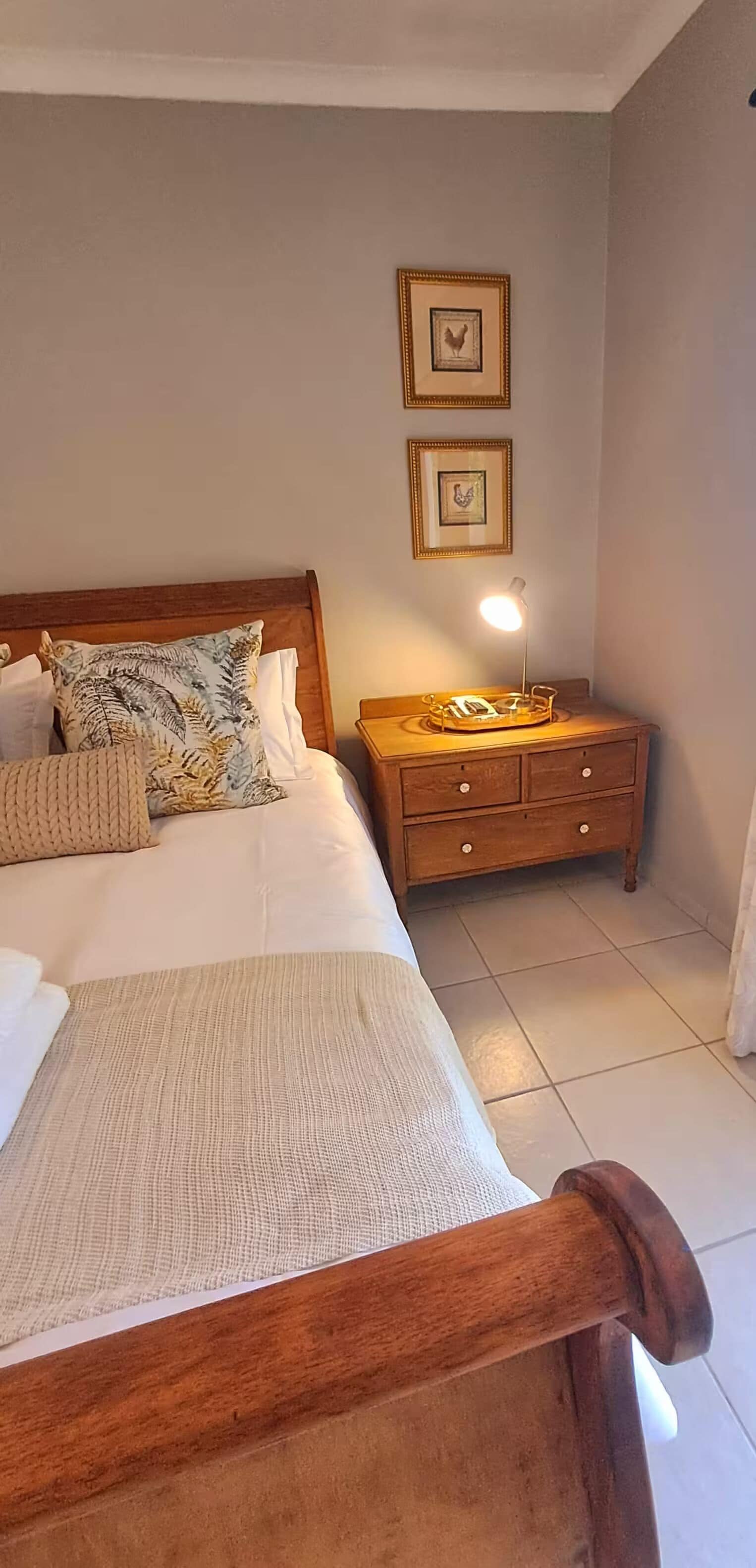 Una cama bien hecha con cabecera de madera, una mesita de noche con lámpara y dos cuadros enmarcados en la pared.