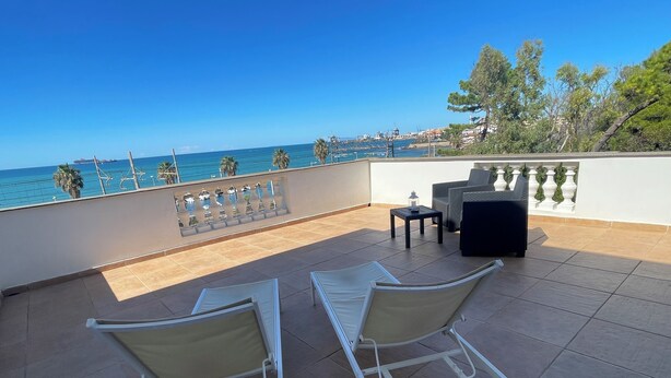 A terrace with two white lounge chairs, a small table, and a view of the sea and buildings in the background.