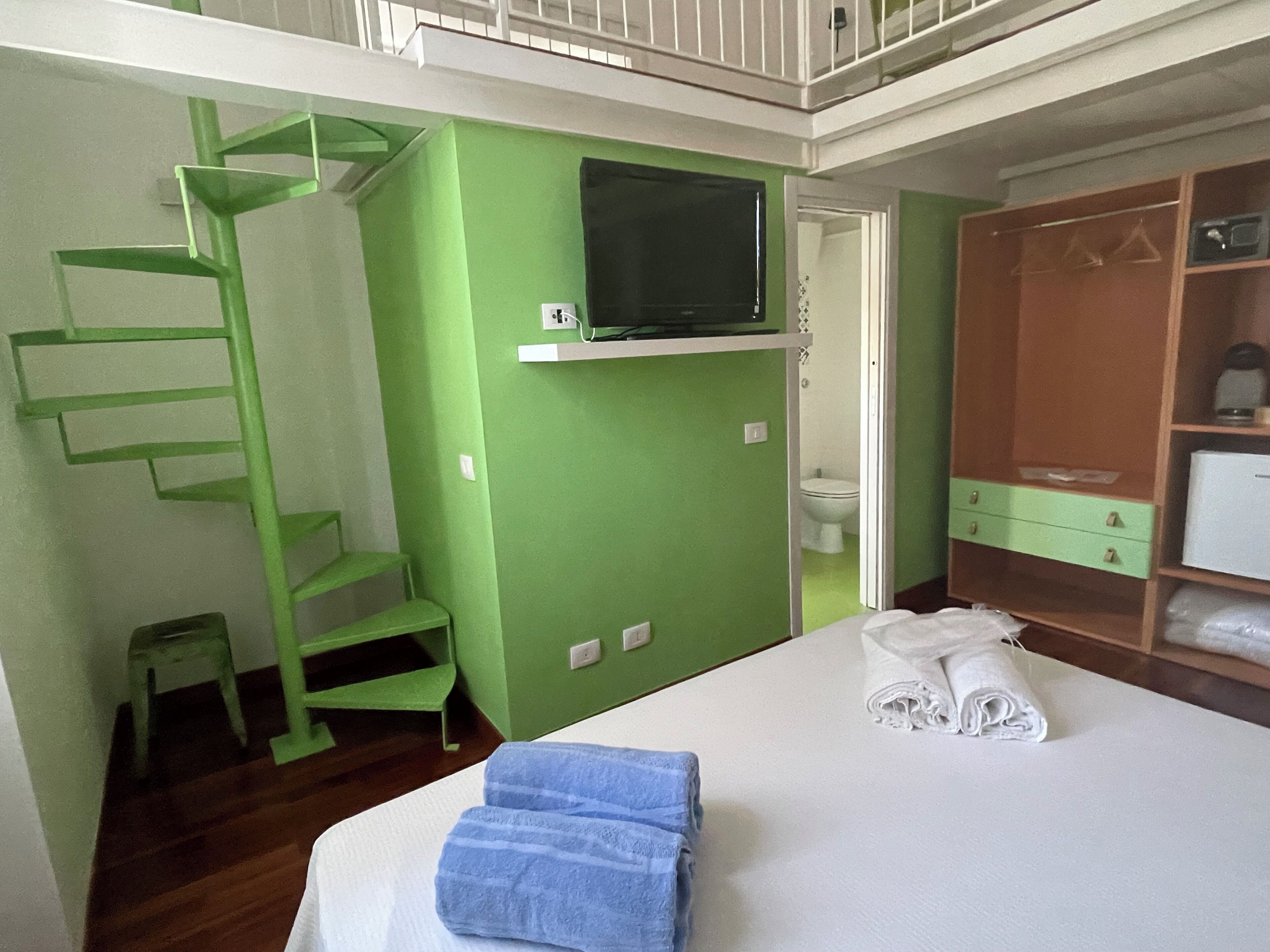 A room with a bed, a green wall, a TV mounted above it, a wooden wardrobe, and a small green staircase.