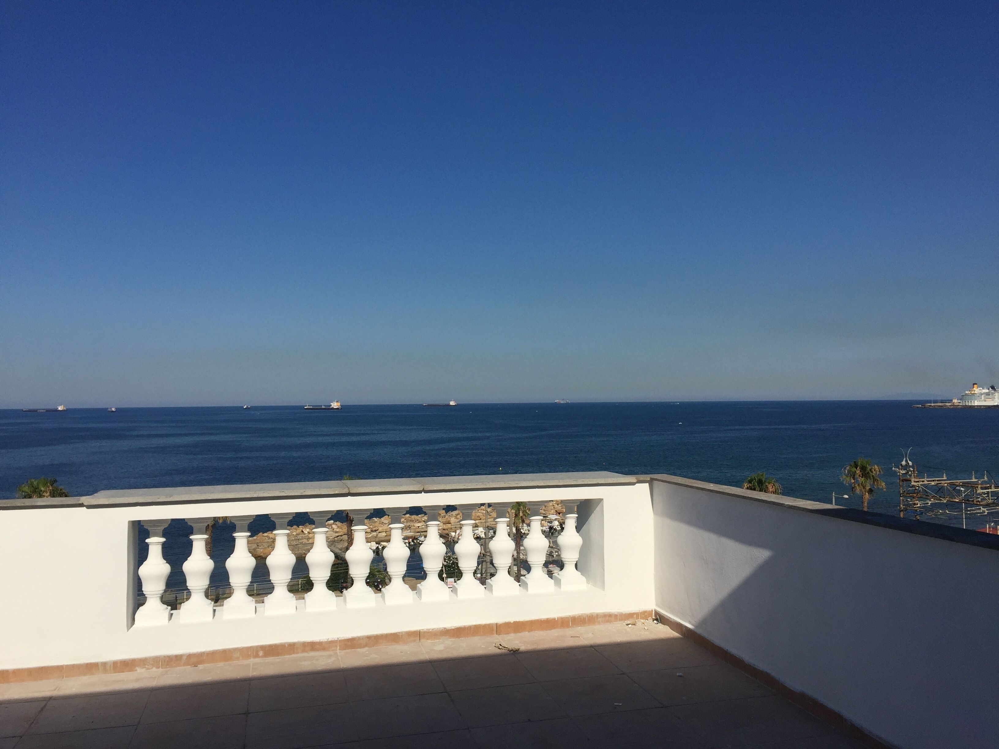 A balcony with a white balustrade overlooking a calm sea with several ships in the distance.