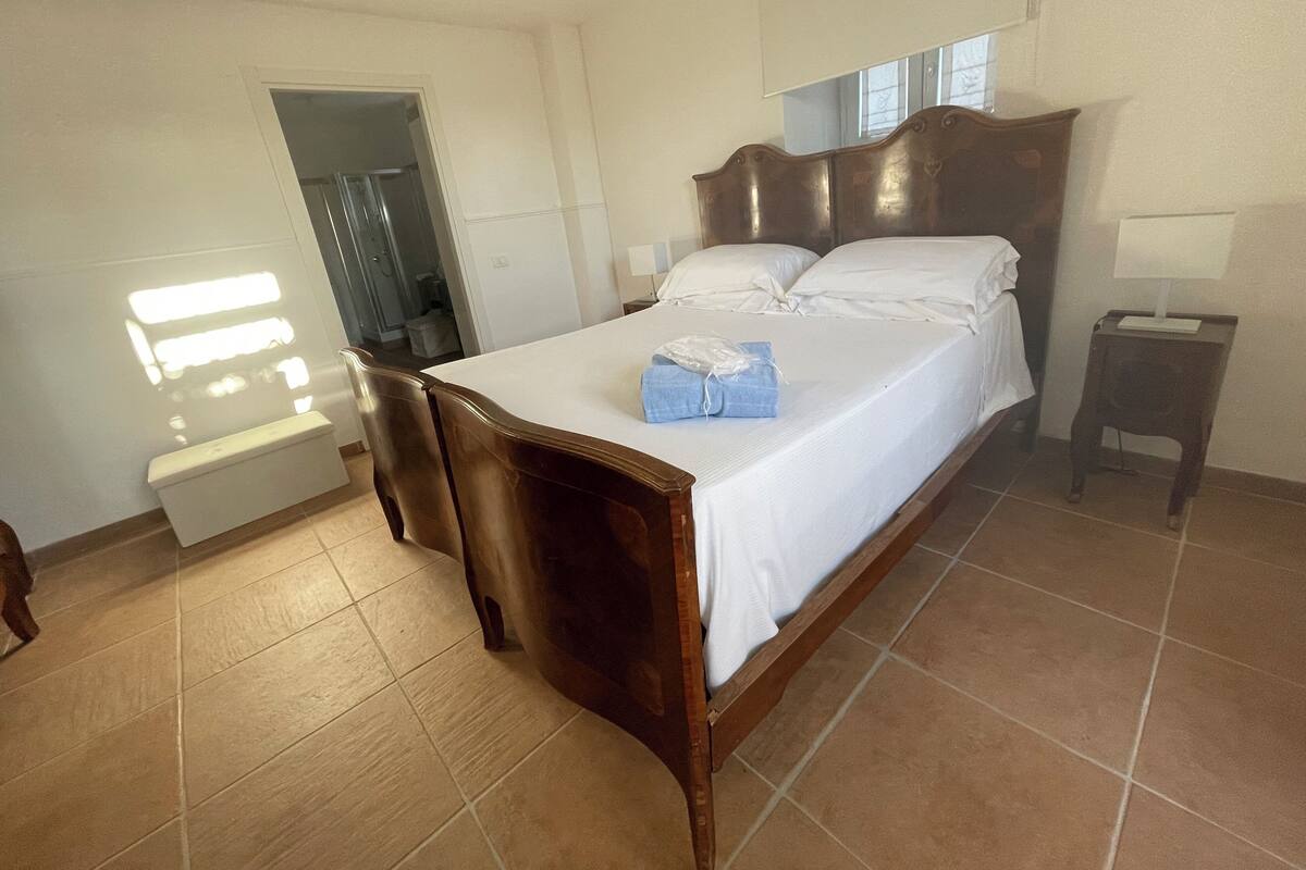 Une chambre à coucher avec un lit en bois, une table de chevet, une commode et une salle de bain visible à travers une porte ouverte.