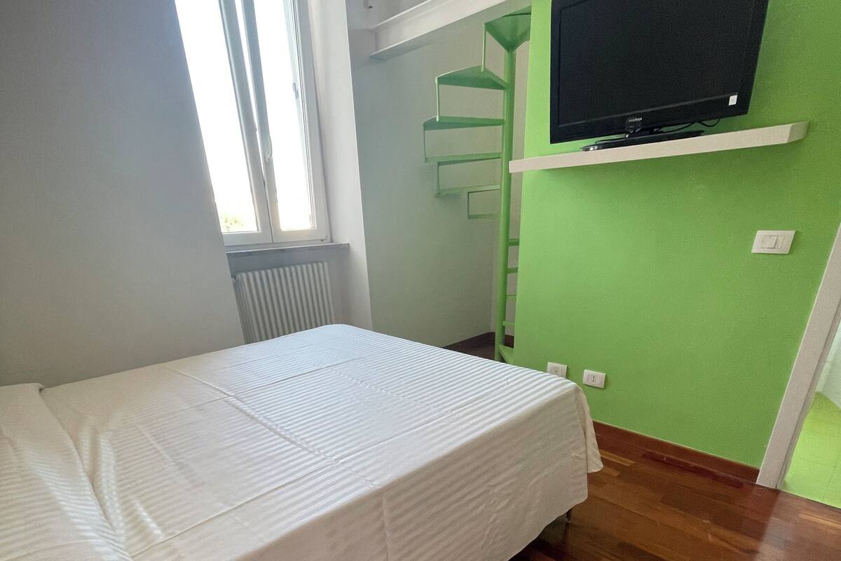 Une chambre avec un lit superposé, un mur vert, un téléviseur fixé au mur et une fenêtre avec des stores.