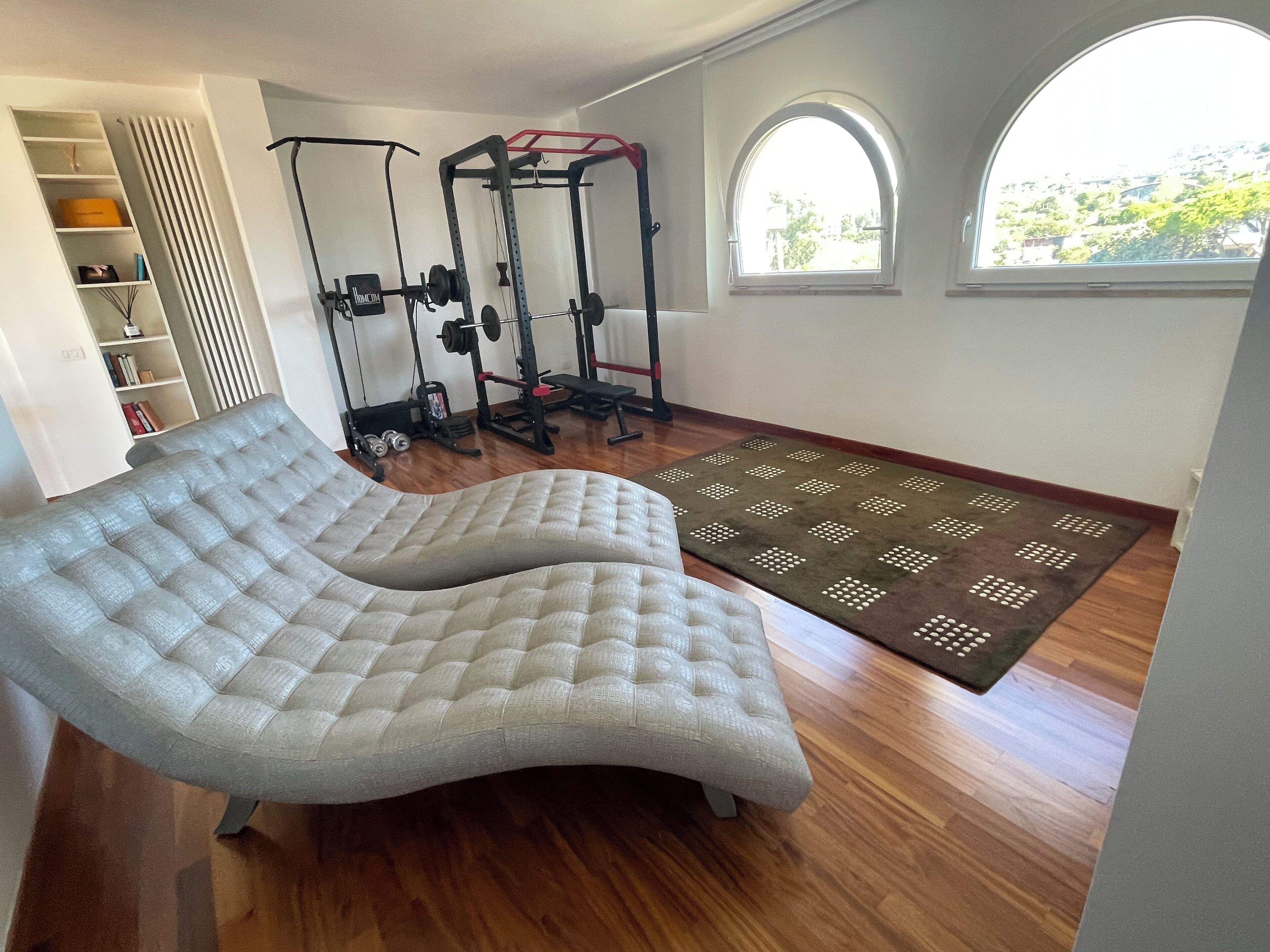 A room with a large cushioned bench, a weightlifting area, and a view through a window.
