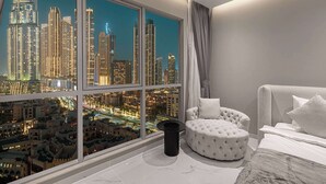 A modern hotel room with a large window offering a city view, a round ottoman, and a neatly made bed.