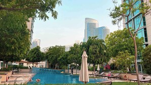 A swimming pool area with lounge chairs and umbrellas, surrounded by trees and modern buildings in the background.