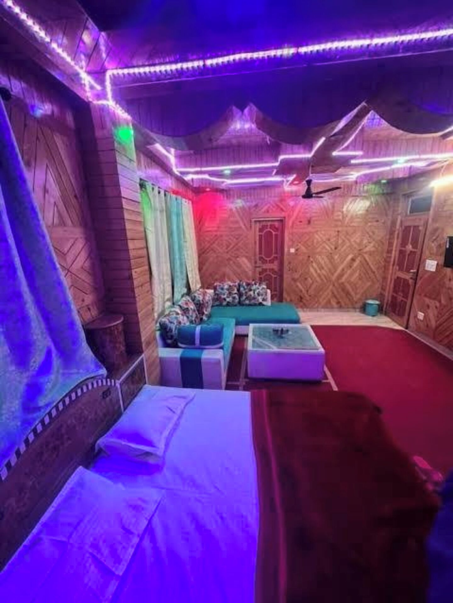 A room with a bed, a sofa, and a small table. There are wooden walls and a ceiling fan.