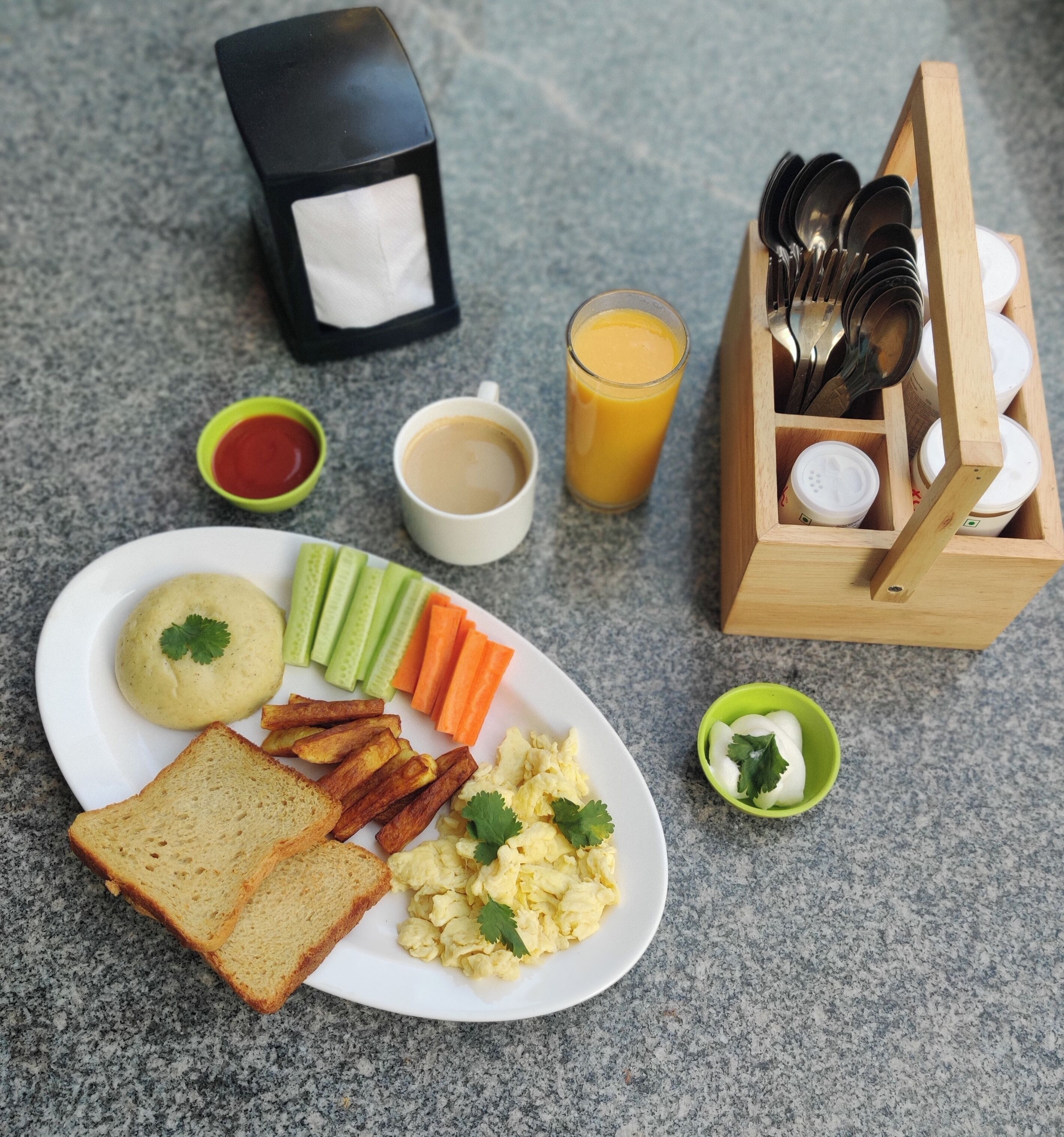 A breakfast plate with toast, scrambled eggs, and vegetables, a glass of orange juice, a cup of coffee, and a box of utensils.