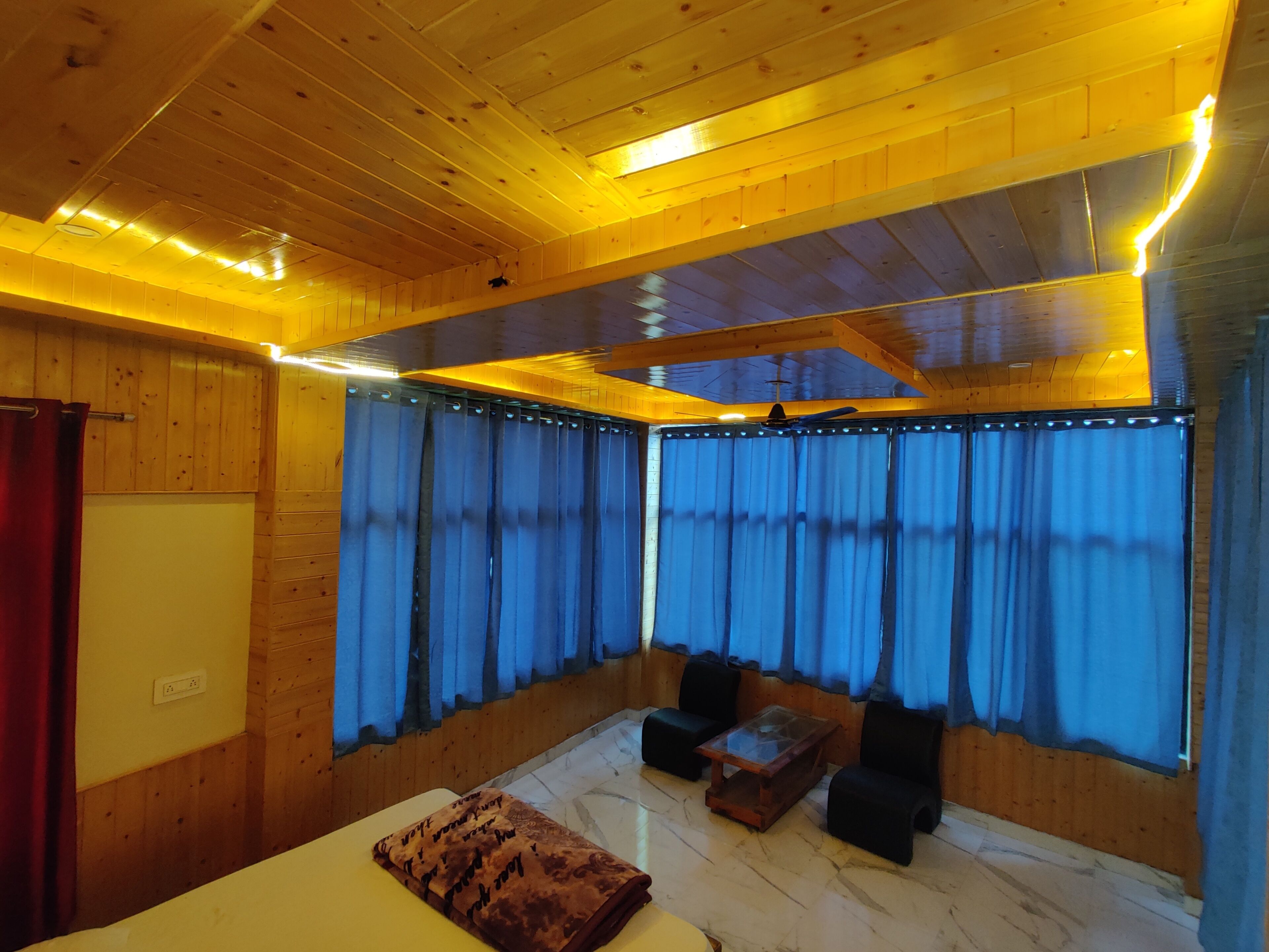 Une pièce aux murs et au plafond en bois, avec un lit, une petite table, deux chaises et une grande fenêtre ornée de rideaux bleus.