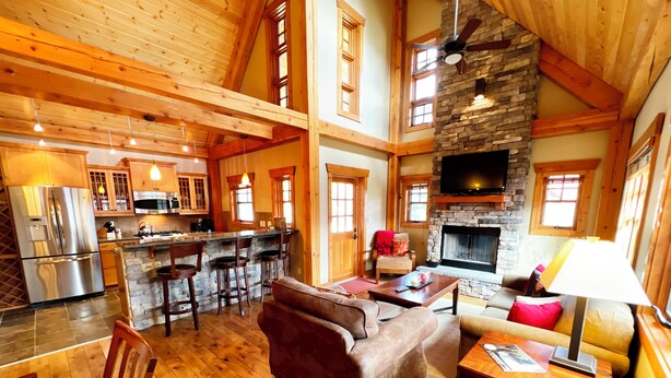 A spacious living area with a stone fireplace, wooden ceiling, and a kitchen with a large island. - Dreamcatcher Mountain Cabin - Ski-Bike-Golf-Relax (Kimberley)