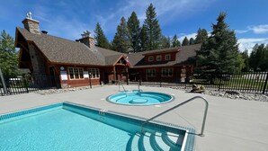 A pool area with a large rectangular pool and a smaller circular pool, surrounded by a wooden cabin-style building with a stone chimney. - Dreamcatcher Mountain Cabin - Ski-Bike-Golf-Relax (Kimberley)