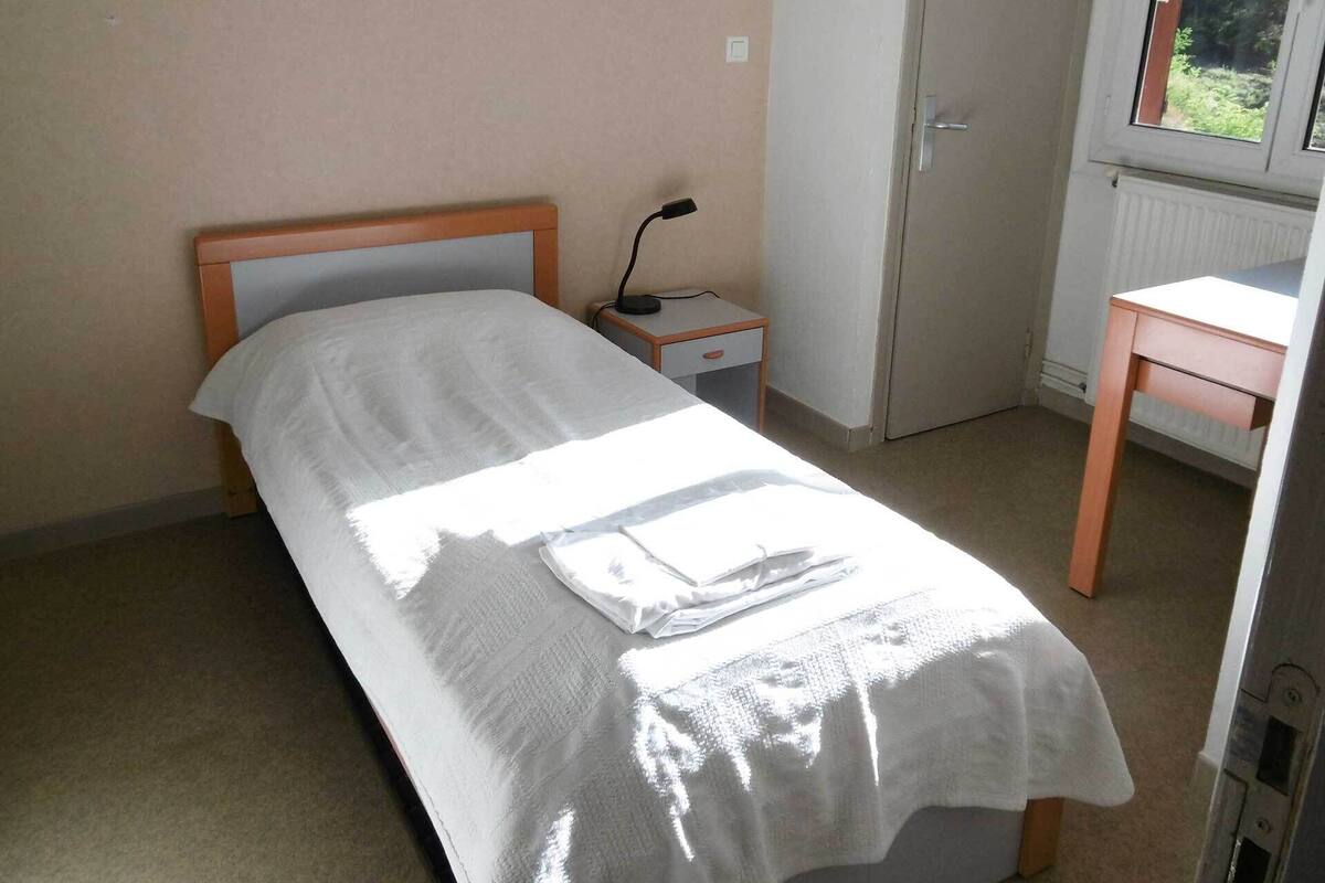 A small, neatly made bed with a white sheet and a folded towel on it. There is a nightstand with a lamp beside the bed, a small wooden desk, and a radiator on the wall.