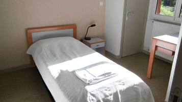 A small, neatly made bed with a white sheet and a folded towel on it. There is a nightstand with a lamp beside the bed, a small wooden desk, and a radiator on the wall.