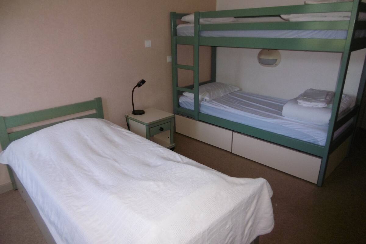 A bunk bed with a single bed below, a bedside table with a lamp, and a small drawer.