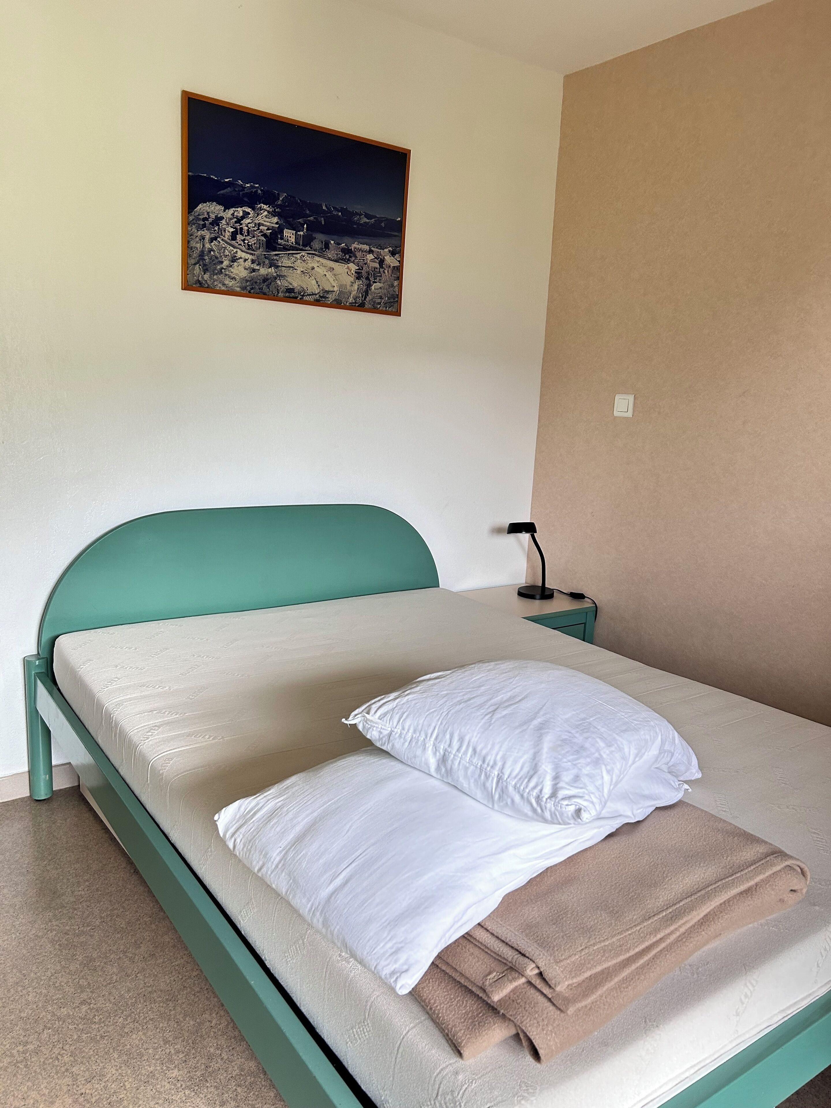 A neatly made bed with a teal headboard, white pillows, and a folded beige blanket. A small bedside table with a lamp is visible.