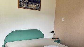A neatly made bed with a teal headboard, white pillows, and a folded beige blanket. A small bedside table with a lamp is visible.