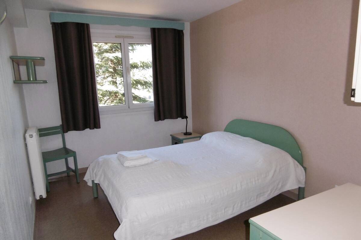 A bedroom with a bed, a chair, a bedside table, and a window with curtains.