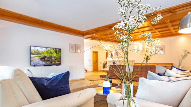 A living room with a sofa, a coffee table, and a vase with flowers. There is a television on the wall and a wooden ceiling. - NEW: Exclusive vacation home at Lechsee in the Allgäu (Lechbruck am See)