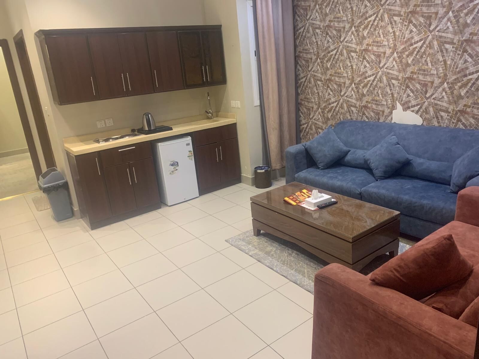 Een compacte woonruimte met een kitchenette, een blauwe bank, een bijzettafeltje en een behang.