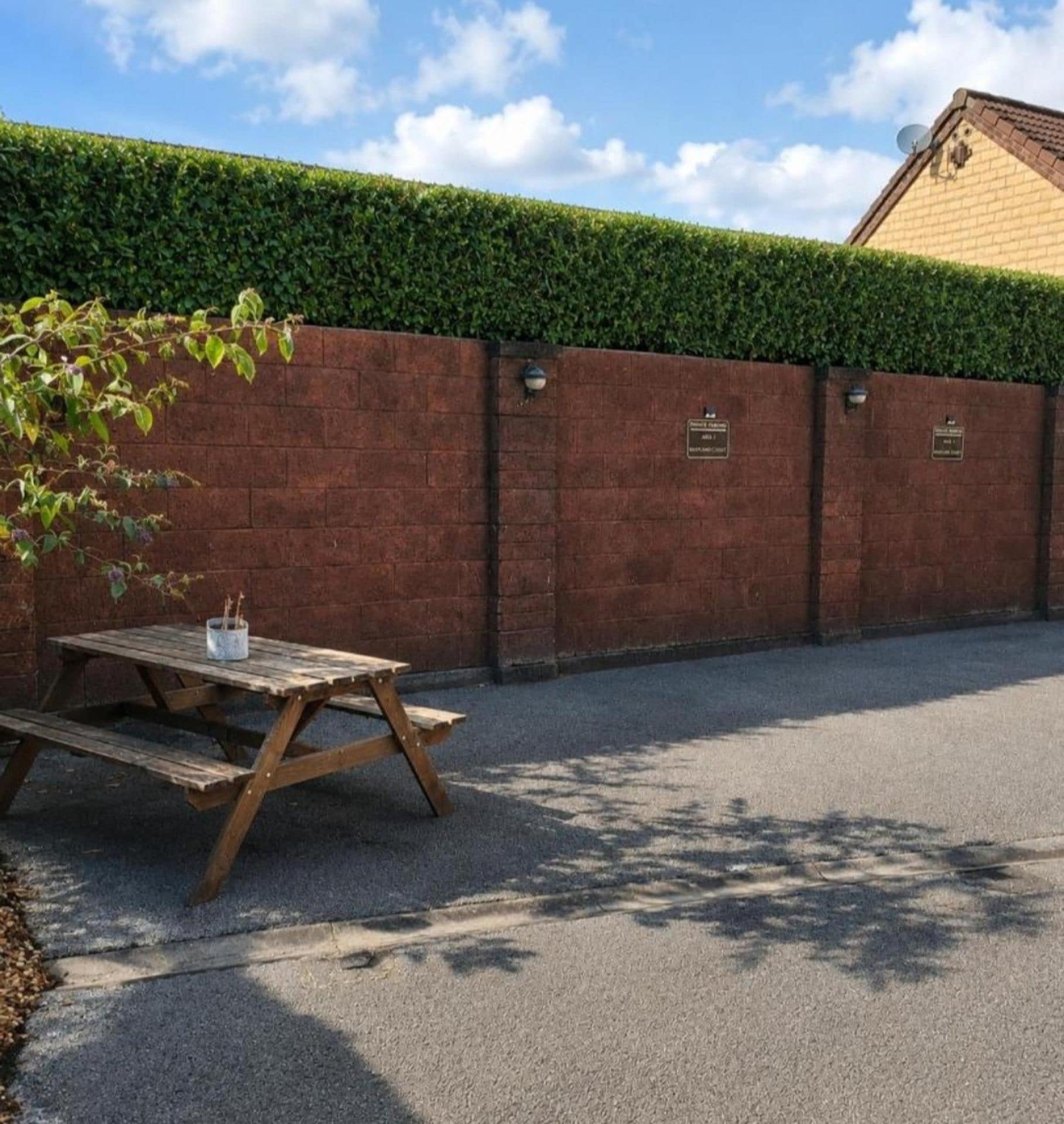 A paved outdoor area with a wooden picnic table and benches, a brick wall with a green hedge, and a building with a brown roof in the background.