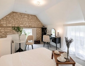 Ein Schlafzimmer mit schrägen Wänden, einer Steinwand, einem Bett, einem Schreibtisch, einem Stuhl, einem Schminktisch und einem Fenster mit transparenten Vorhängen.