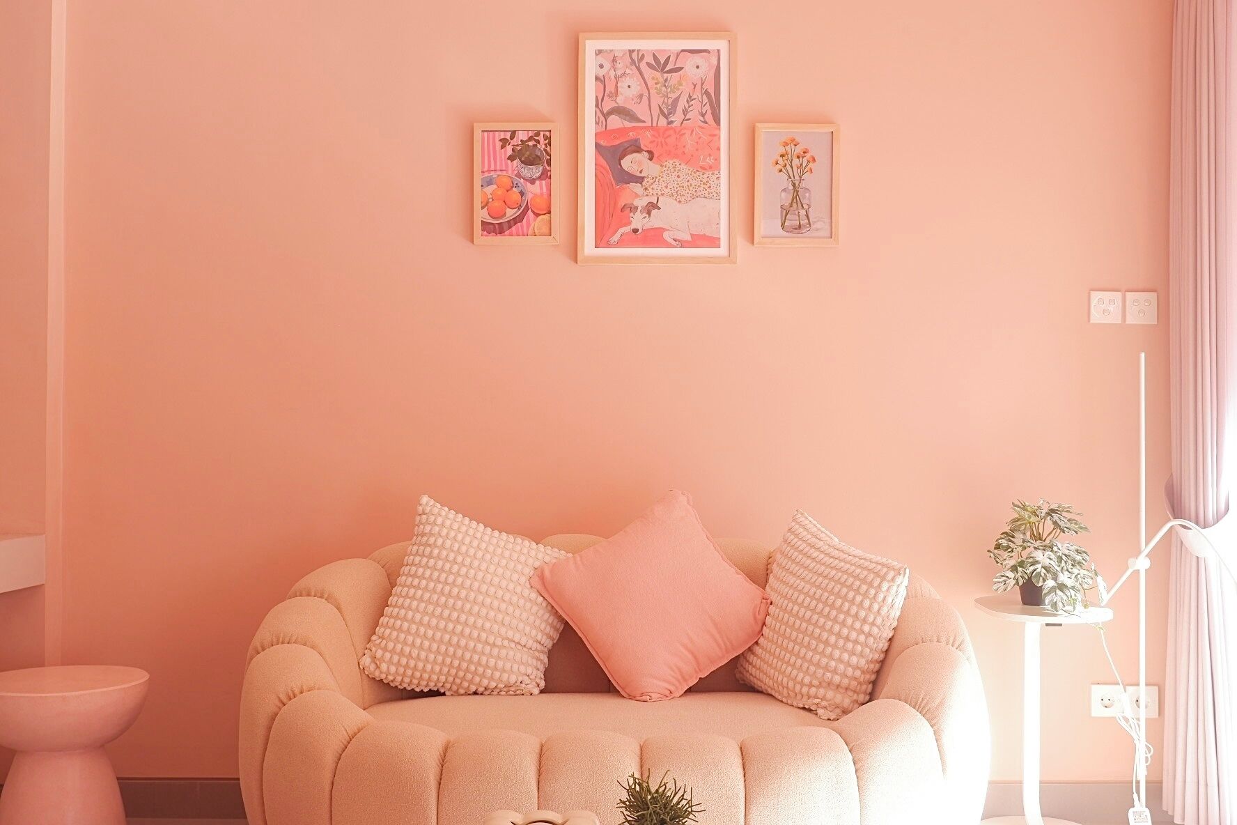 Una stanza accogliente con una parete color pesca, un divano beige rotondo con cuscini rosa e bianchi, e tre opere d'arte incorniciate sopra il divano.